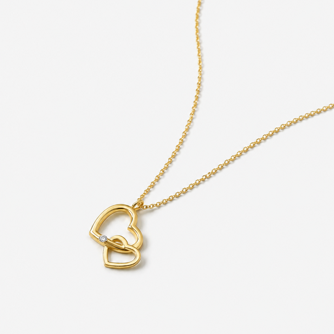 A gold chain necklace featuring two interlocking open heart pendants, one of which has a small clear stone accent, shown on a light gray background.
